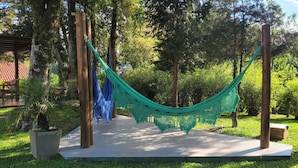 A hammock suspended between two wooden posts in a garden with a concrete pathway, surrounded by greenery and trees. - Guesthouse on Rua Torta, 2 bedrooms, swimming pool, fireplace, free parking. (Gramado)