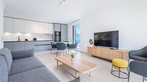 Una sala de estar moderna con sofá, mesa de centro y televisor. Se observa una zona de cocina con armarios y una mesa de comedor.