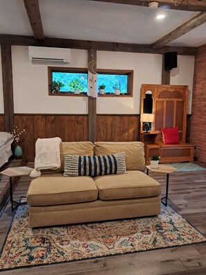 A cozy living room with a beige sofa, a wooden accent wall, a small table with a lamp, and a window with plants. - Forest Nature Retreat with Woodstove and near Durham trails and Thermea Spa (Uxbridge)