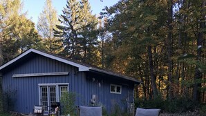 A rustic cabin with a blue exterior, two chairs in front, and a pile of firewood. - Forest Nature Retreat with Firepit Near Durham trails and Thermea Spa (Uxbridge)