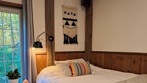 A bedroom with a bed, a bedside table with a plant, a hanging wall decoration, and a window with curtains. - Forest Nature Retreat with Firepit Near Durham trails and Thermea Spa (Uxbridge)