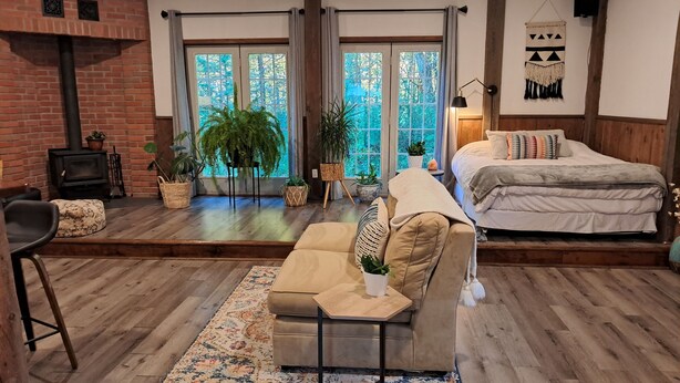 A cozy living room with a fireplace, a sofa, a bed, and large windows with a view of greenery. - Forest Nature Retreat with Firepit Near Durham trails and Thermea Spa (Uxbridge)