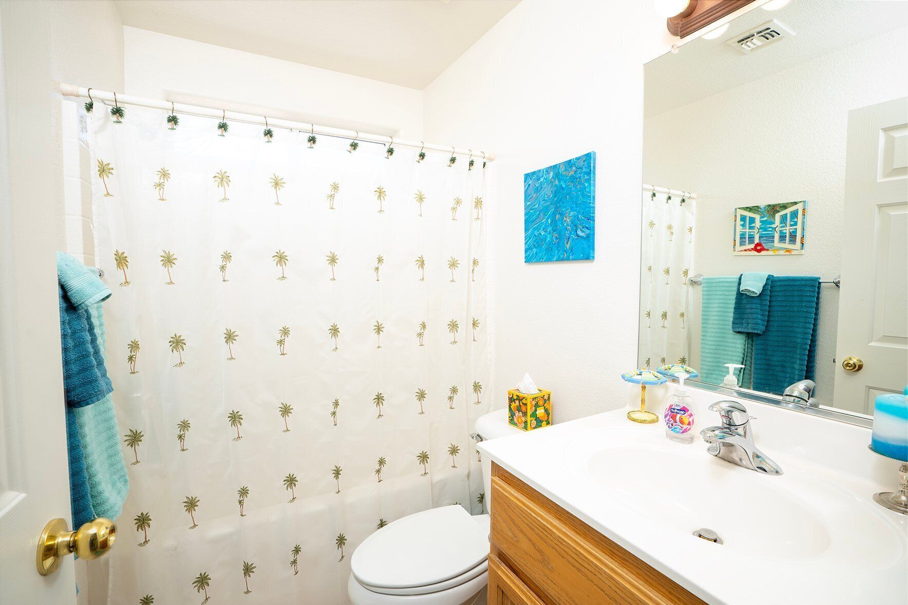 Une salle de bain avec un rideau de douche orné de motifs de palmiers, des toilettes, un lavabo avec un miroir et un porte-serviettes sur lequel est posée une serviette bleue.