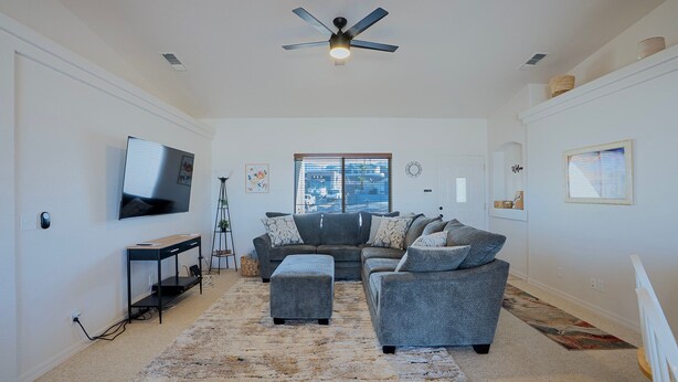 Ein geräumiges Wohnzimmer mit Deckenventilator, großem Flachbildfernseher, grauem Sofasatz und Fenster mit Jalousien.