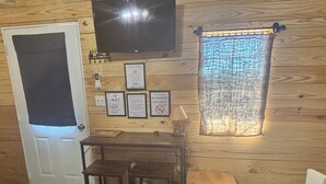 Una habitación pequeña y acogedora con paredes de madera, un televisor montado en la pared, un escritorio con dos sillas, una ventana con cortina y una puerta con luz encima.