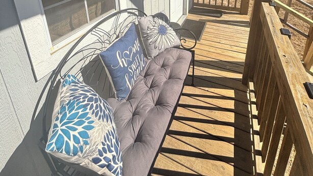 Un porche cubierto con un banco acolchado y almohadones decorativos, baranda de madera y vistas despejadas del paisaje circundante.