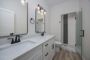 Une salle de bain avec un meuble-lavabo blanc, un robinet noir, un grand miroir avec éclairage intégré, des toilettes et une douche à parois de verre.