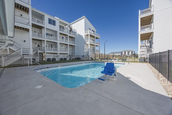 Une piscine avec une chaise bleue, entourée d’une terrasse en béton et d’un bâtiment blanc doté de balcons.