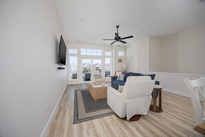 A spacious living room with a flat-screen TV, a ceiling fan, and a large window offering a view of the city.
