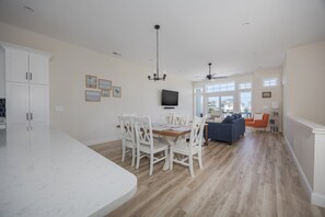 Una cocina amplia con comedor, una sala de estar con sofá y televisor, y vistas al exterior a través de amplias ventanas.