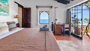 Un dormitorio con una cama grande, un escritorio y vista al océano a través de una ventana.
