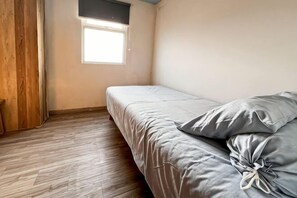 A single bed with a grey mattress and pillows, a wooden floor, a window with blinds, and a plain wall. - BEST View 2BR Apt Pluit w/ Pool WiFi & Netflix (North Jakarta)