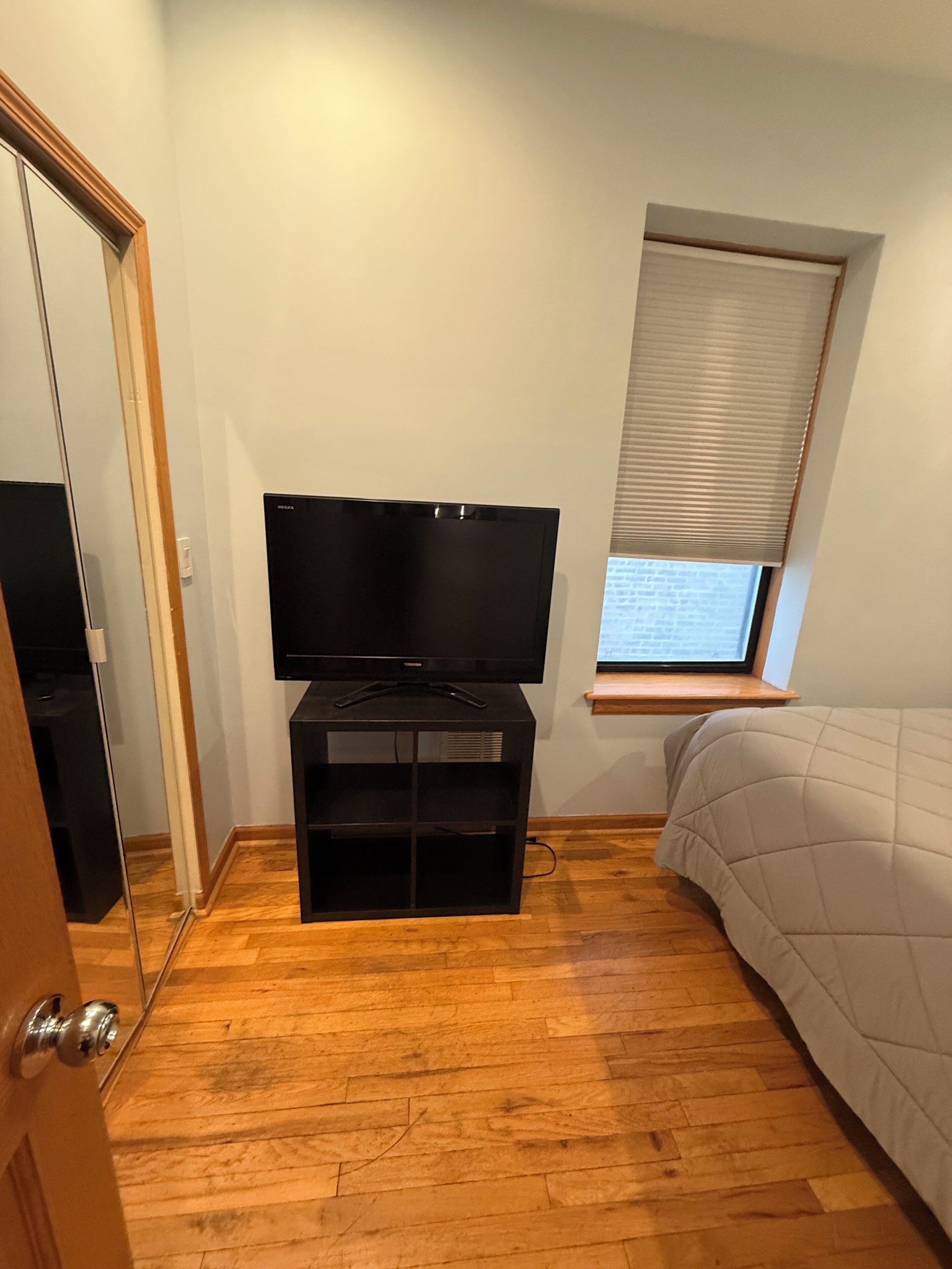 Un dormitorio con televisor sobre un soporte, una ventana con persianas, una cama con cubrecama acolchado y suelo de madera.
