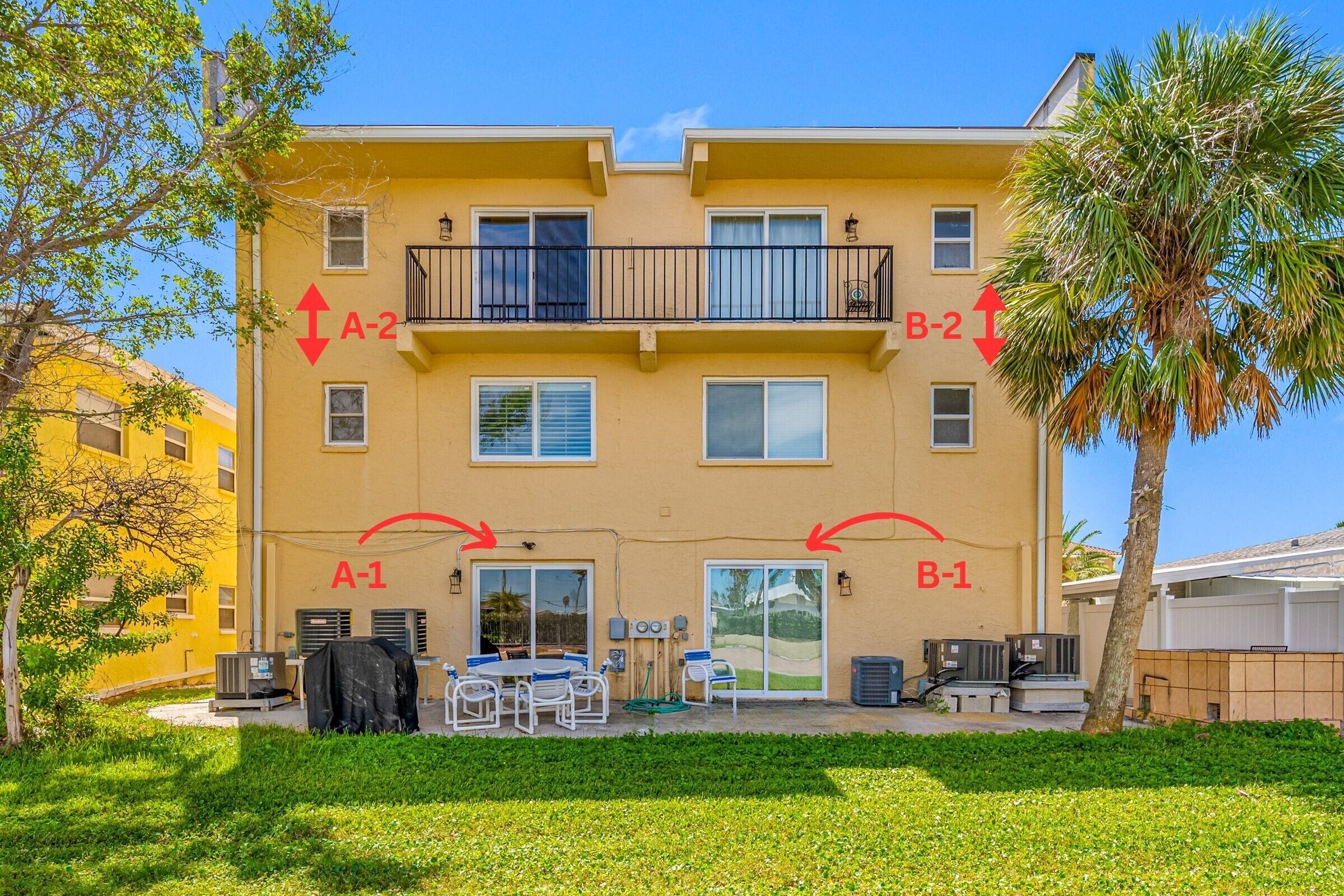 A two-story apartment building with balconies, a covered patio with outdoor furniture, and a palm tree.