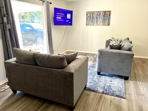 A living room with a sofa and armchair, a flat-screen TV, and a painting on the wall.
