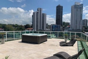A rooftop terrace with a hot tub, lounge chairs, and a glass balustrade overlooking a cityscape with tall buildings.
