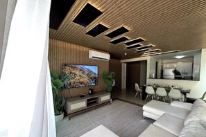 Una sala moderna con televisor de pantalla plana, un comedor con sillas y una cocina visible a través de un umbral abierto.