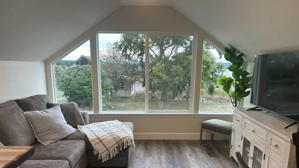 A cozy living room with a gray sofa, a bench, and a TV on a wooden cabinet. Large windows offer a view of the outdoors.
