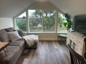 A cozy living room with a gray sofa, a bench, and a TV on a wooden cabinet. Large windows offer a view of the outdoors.