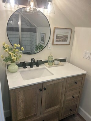 A bathroom with a round mirror, a white sink, wooden cabinets, and a potted plant.