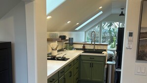 A modern kitchen with green cabinets, a stainless steel sink, and a built-in oven. There is a window providing natural light and a view of the outdoors.