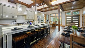 A spacious kitchen with wooden beams, a long island with stools, and a dining area with a table set for four. - Panorama Peak | 8BR Luxury Lodge w/ Pickleball, Game Room & Stunning Mountain Views (Broken Bow)