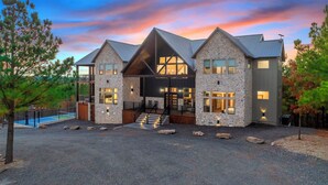 A modern two-story building with a stone facade, a covered entrance, and a pool area. - Panorama Peak | 8BR Luxury Lodge w/ Pickleball, Game Room & Stunning Mountain Views (Broken Bow)