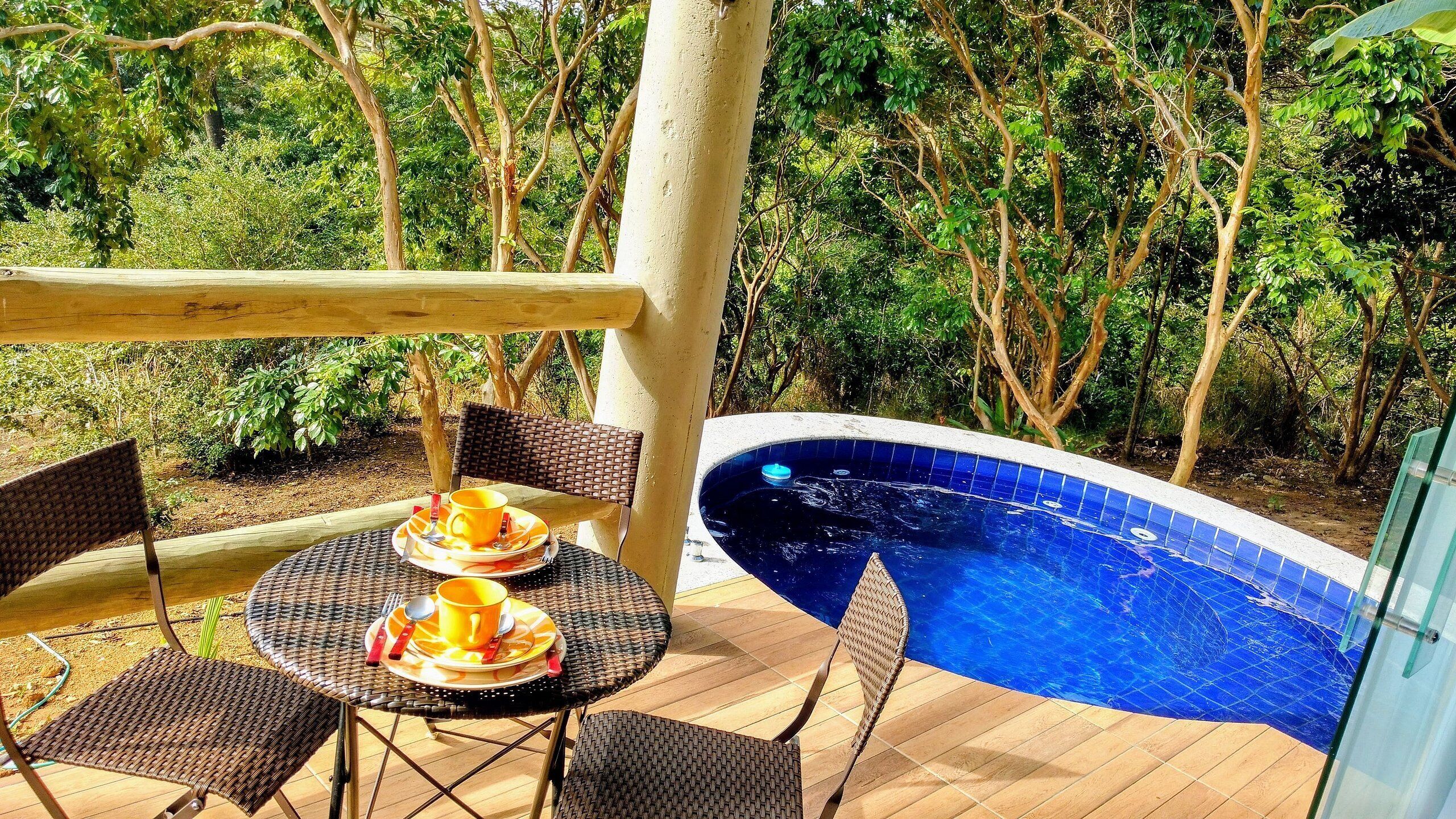 Ein Sitzbereich im Freien mit Korbstühlen und einem Tisch, ein kleiner Pool mit blauen Fliesen und üppiges Grün im Hintergrund.