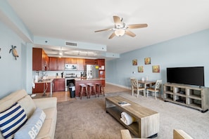 A living room with a sofa, coffee table, TV stand, and a kitchen area with a bar and stools. - Spacious East PCB Condo w/ Lake Views, Pools & Easy Beach Access (Panama City Beach)