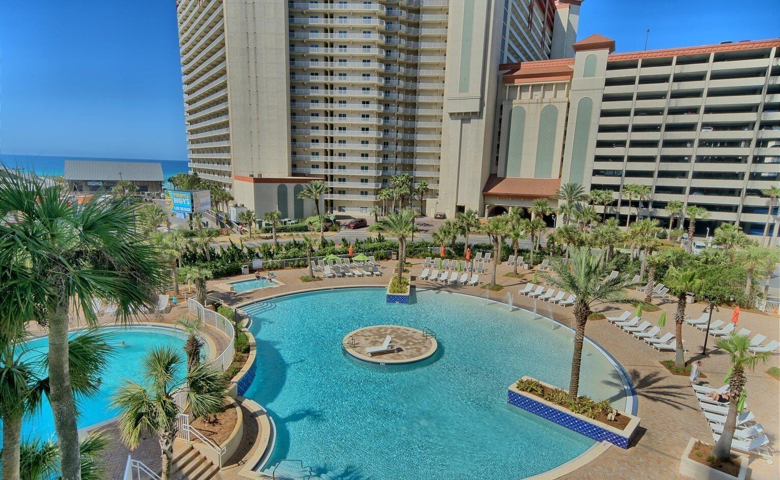 A hotel with a large central pool surrounded by lounge chairs and palm trees.
