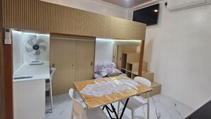 Una habitación compacta con techo de madera, un escritorio blanco con ventilador, una mesa con mantel de encaje y una cama litera con manta estampada.