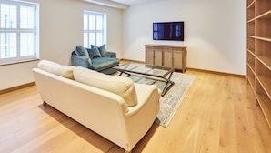 A living room with a beige sofa, a glass coffee table, a blue sofa, a wooden cabinet, and a TV on the wall.