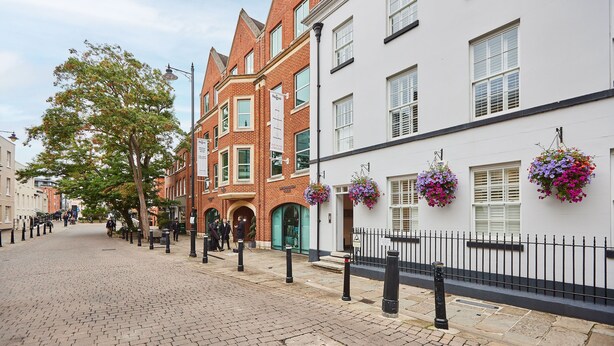 A cobblestone street lined with traditional brick and white buildings, some with flower baskets.