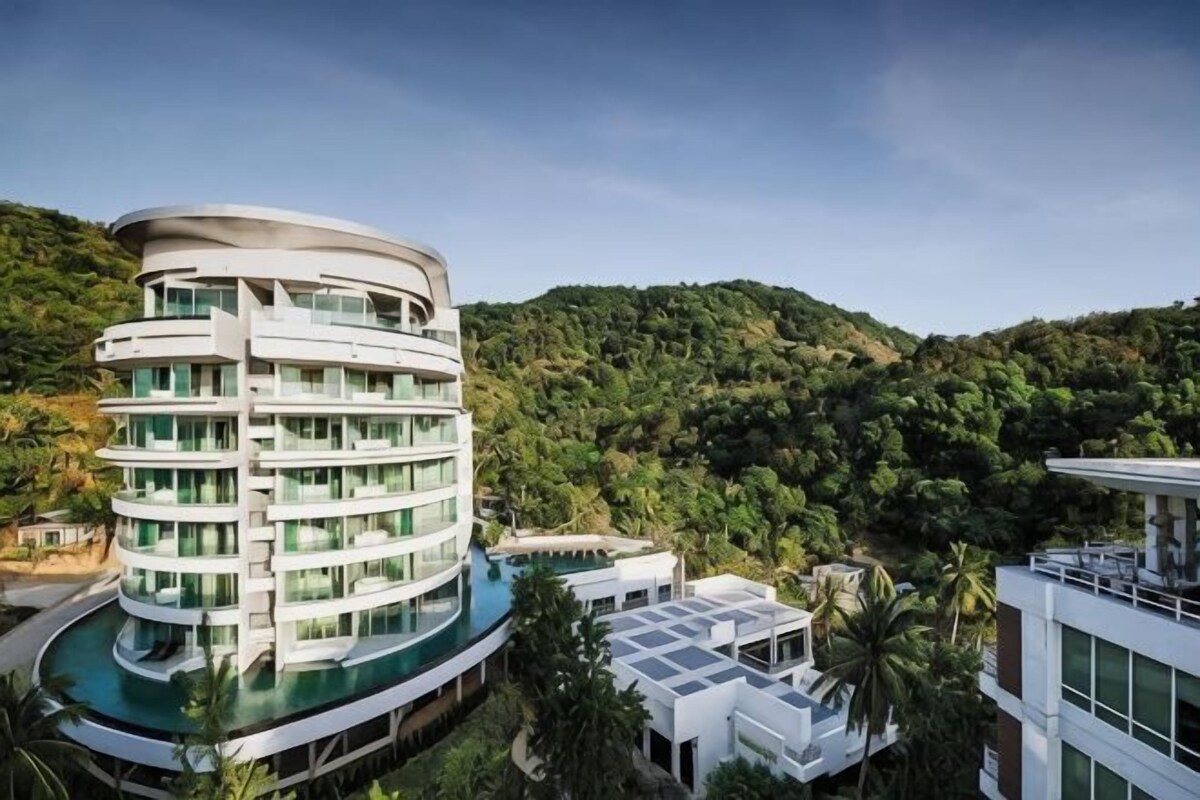 A multi-story hotel with a curved pool, surrounded by lush greenery and hills.