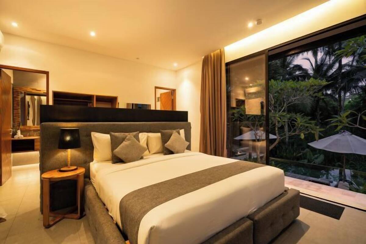A modern bedroom with a large bed, a nightstand, a mirror, and a view of a tropical garden through a glass door.