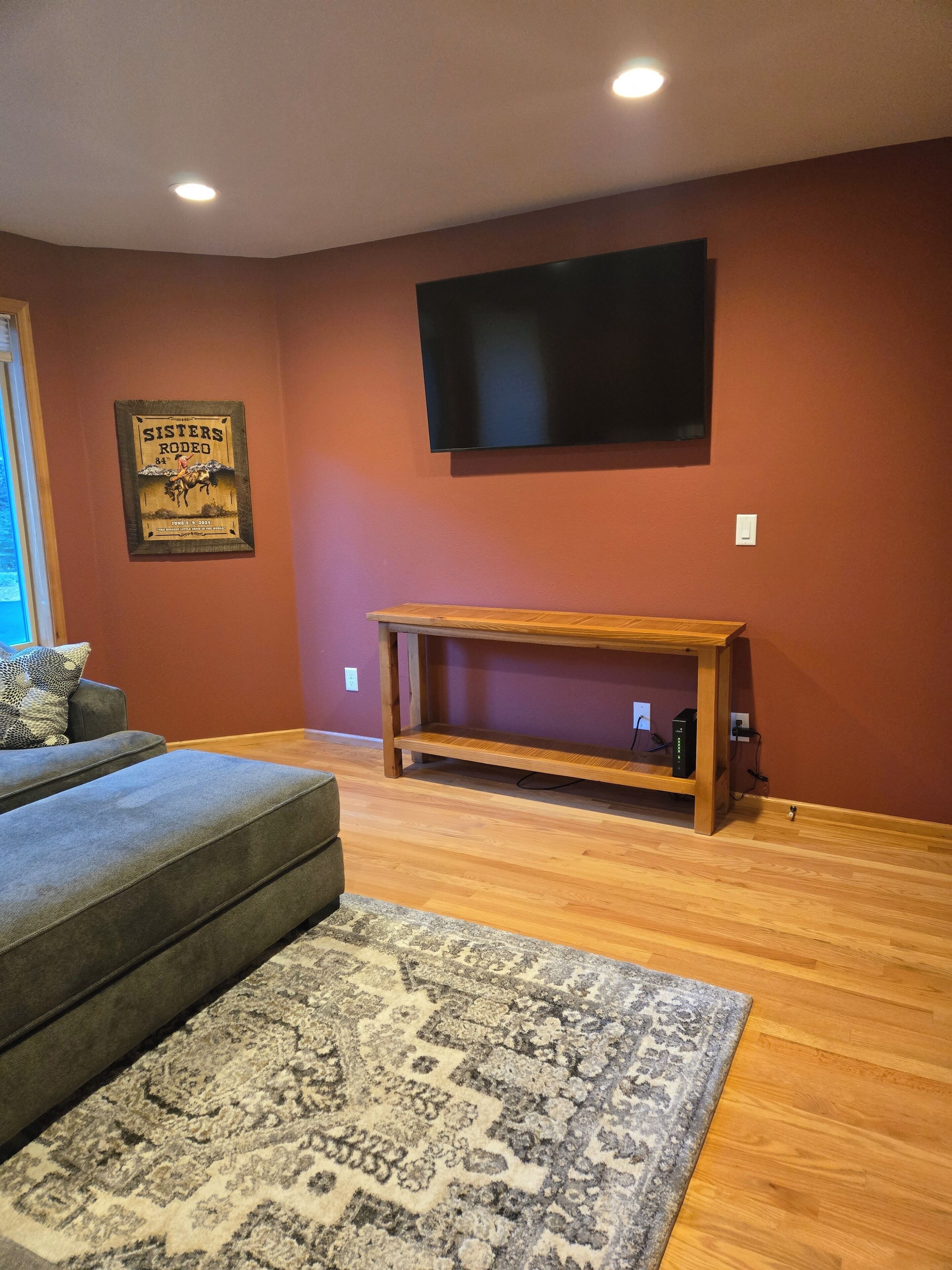 Una sala de estar con un televisor de pantalla plana montado en la pared, un mueble de televisión de madera, un sofá gris y una alfombra estampada.