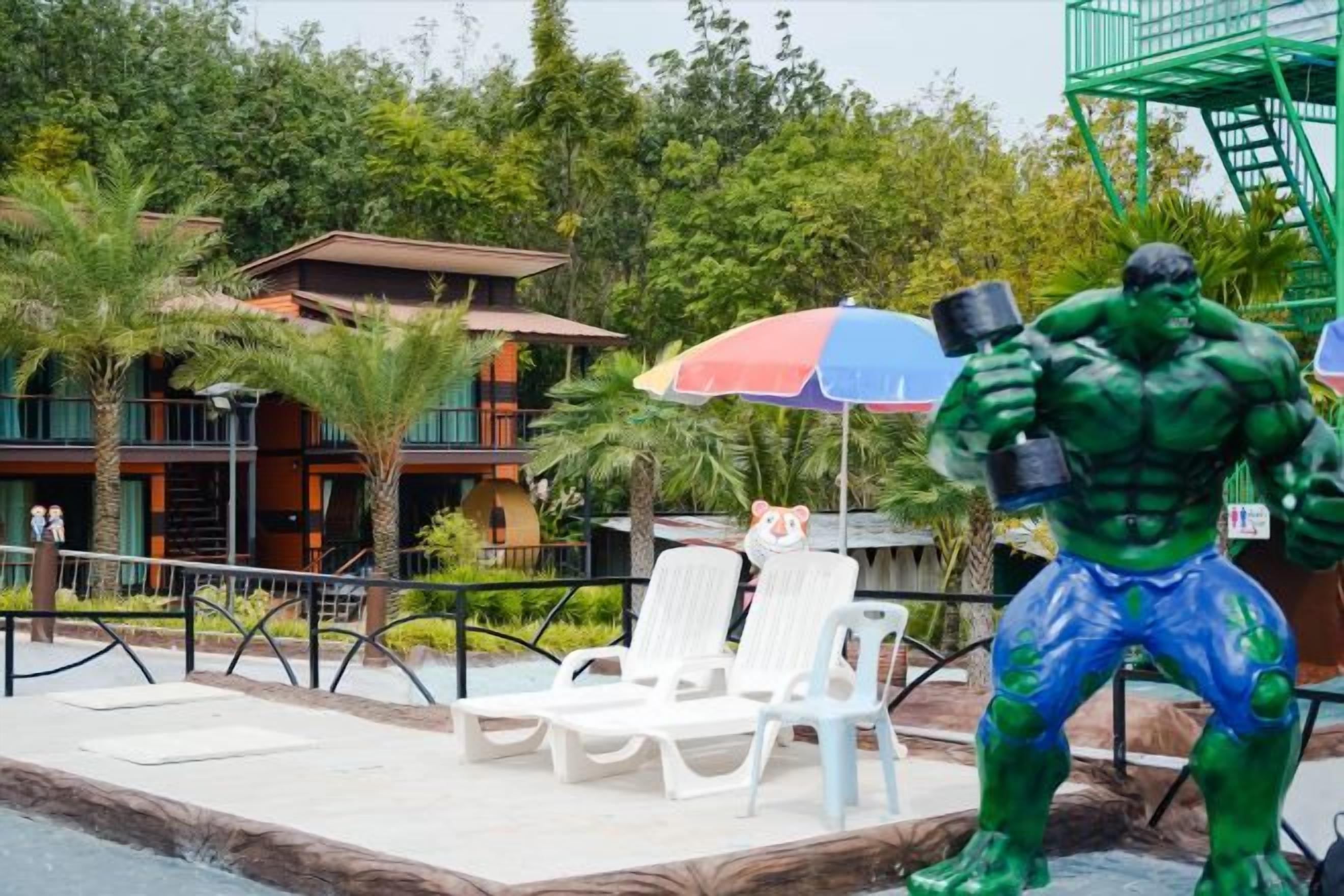 Eine Statue einer muskulösen Figur mit einem Hammer, ein Poolbereich mit Liegestühlen und ein Gebäude mit Balkonen im Hintergrund.