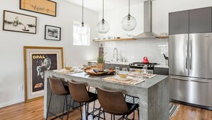 Een moderne keuken met een centraal eiland, roestvrijstalen apparatuur, hanglampen en lijstwerken aan de wanden.