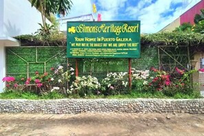 A sign for Simon's Heritage Resort with a green background and white text, surrounded by a garden with pink and white flowers. - Simons Heritage Resort Puerto Galera (Puerto Galera)