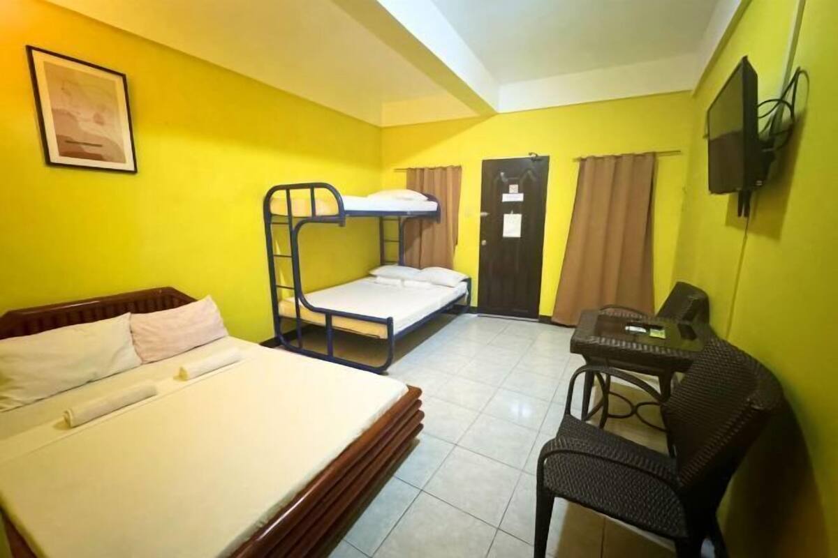 A room with a bunk bed, a single bed, a desk with a chair, and a television mounted on the wall.