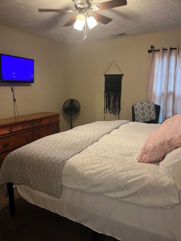 A bedroom with a bed, a dresser, a ceiling fan, a wall-mounted TV, a fan, and a patterned wall hanging. - Cozy 3-bedroom cottage in Hope Mills (Hope Mills)