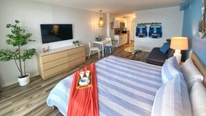 A modern hotel room with a large bed, a desk, a dining area, and a kitchenette.