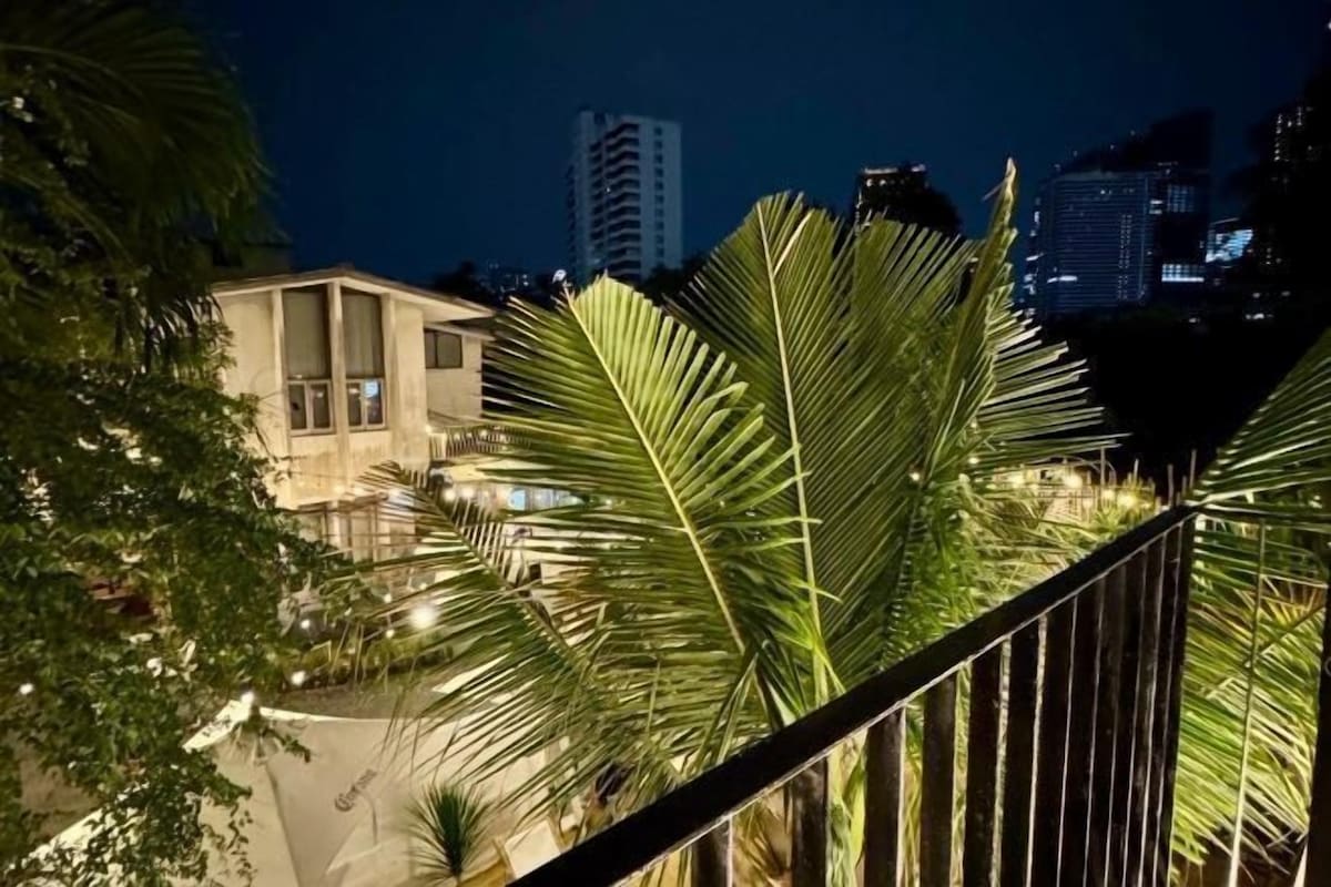 A nighttime view of a building with a balcony, palm trees, and city lights in the background.
