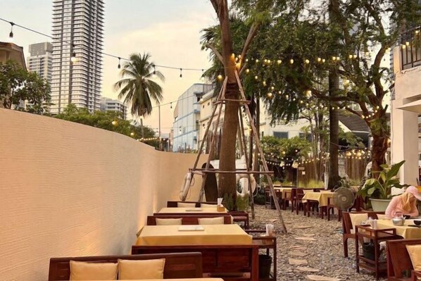 Un área de comedor al aire libre con mesas, sillas, luces colgantes y un árbol.