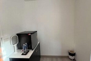 A small room with a desk, a fan, two water bottles, a trash bin, and a heater.