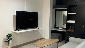 Een moderne woonkamer met een flatscreen televisie, een houten salontafel, een bank, een kleine plant en inbouwplanken met handdoeken.