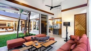 A modern living room with a large sofa, a coffee table, and a dining area. There is a ceiling fan, a lamp, and a view of the outdoor garden. - Villa Crazy Roof by BaliSuperHost (Bali)