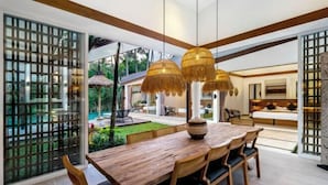 A modern dining area with a wooden table and chairs, a woven basket centerpiece, and a view of the outdoor garden through large glass doors. - Villa Crazy Roof by BaliSuperHost (Bali)