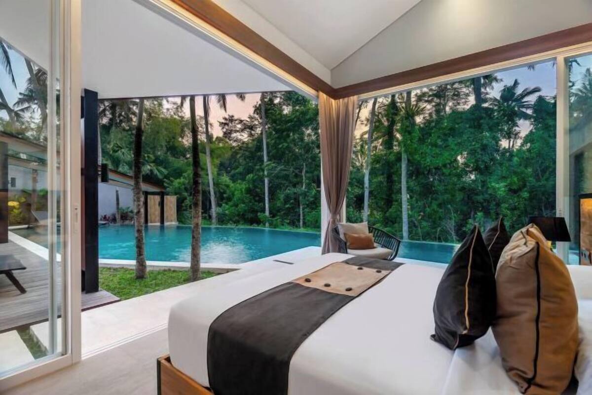 A bedroom with a large window, a bed, and a view of a swimming pool and greenery.