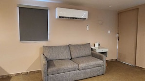 Ein Zimmer mit grauem Sofa, einem kleinen weißen Beistelltisch und einem Fenster mit Jalousien.